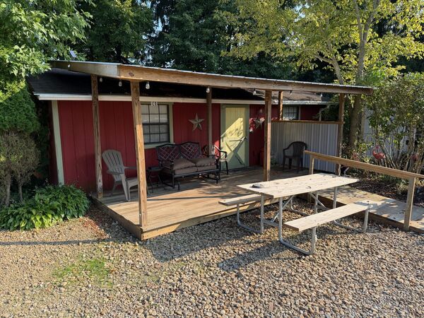Covered porch of the red bunkhouse with a cushioned loveseat, an Adirondack chair and other seating, and a picnic table on the gravel in front