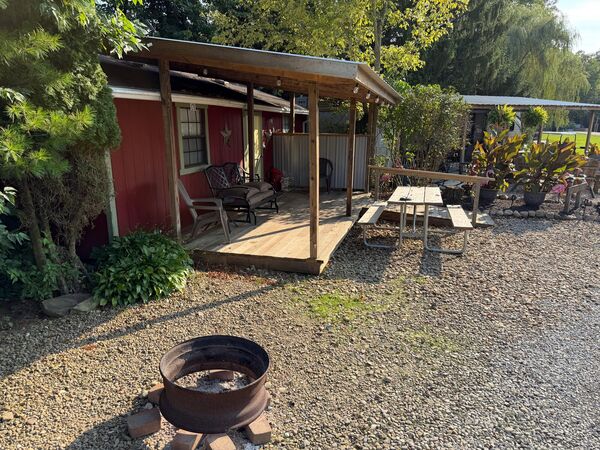 Red bunkhouse from the side, its covered porch and seating behind a picnic table, with a fire ring in the foreground and potted plants nearby