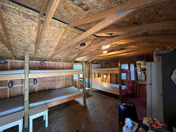 Long open room with numbered wooden bunk beds along both walls, small stools beside them and a full-size refrigerator at the far end
