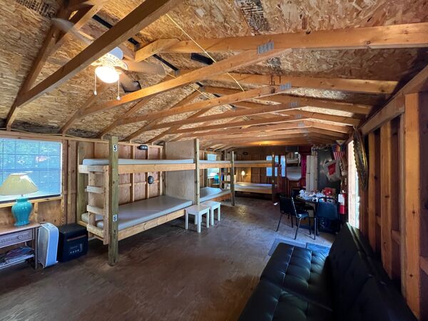 Numbered wooden bunk beds and stools down the room, a table lamp by the window, and a table with chairs and a couch at the far end