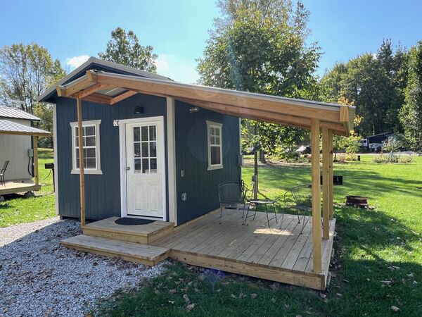 New wooden deck with a slanted roof beside the front door, holding two metal chairs and a small table, with a fire ring on the lawn