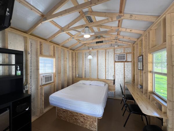 Queen bed on a platform under a ceiling fan, with wall air conditioners, a black storage shelf and a long wooden counter with two chairs under a window