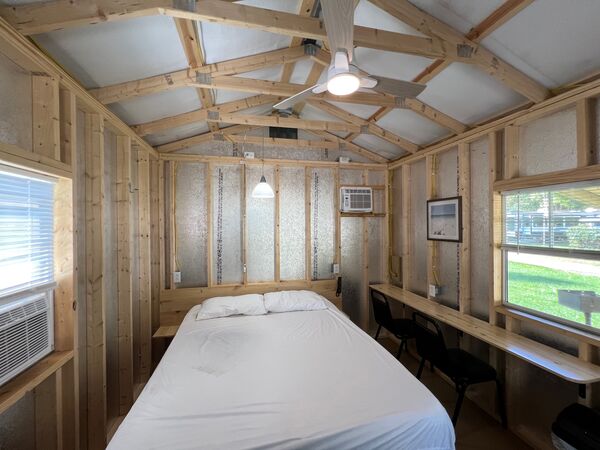 Queen bed beneath a vaulted ceiling with exposed rafters and a fan, a long counter with two chairs along the window wall