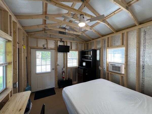 Front door and mounted television seen past the bed, with a black shelf unit holding a mini fridge and microwave, and a window air conditioner