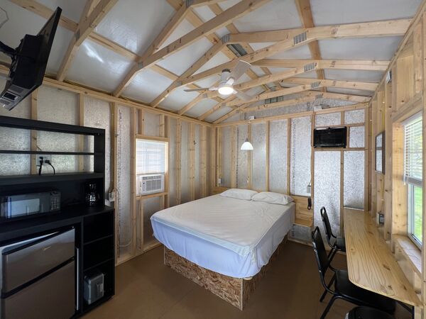 Queen bed under a vaulted ceiling with a fan, a television on a wall arm, a black shelf with a microwave and mini fridge, and a long counter with chairs