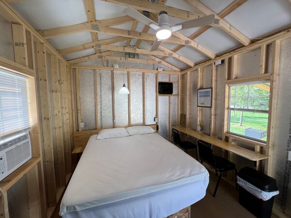 Queen bed with two pillows beneath a pendant light, a window air conditioner on one side and a long counter with chairs under the window on the other