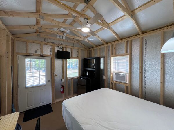 From the bed toward the front door: a mounted television, a black shelf unit with a mini fridge, and a window air conditioner