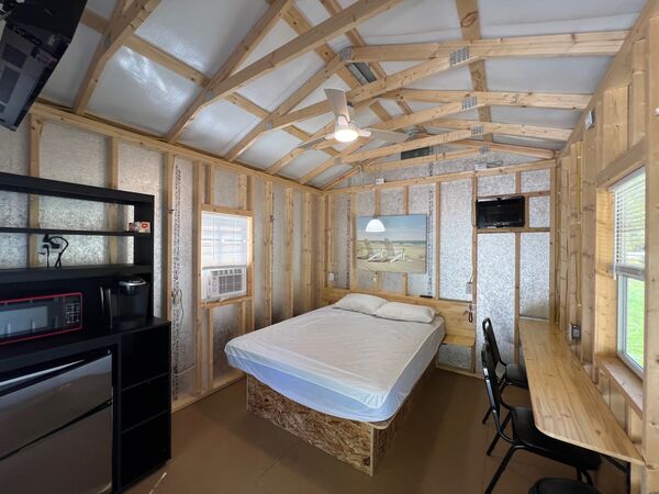 Queen bed beneath a beach-chair print and pendant light, with a black shelf unit holding a microwave, coffee maker and mini fridge, and a long counter with chairs