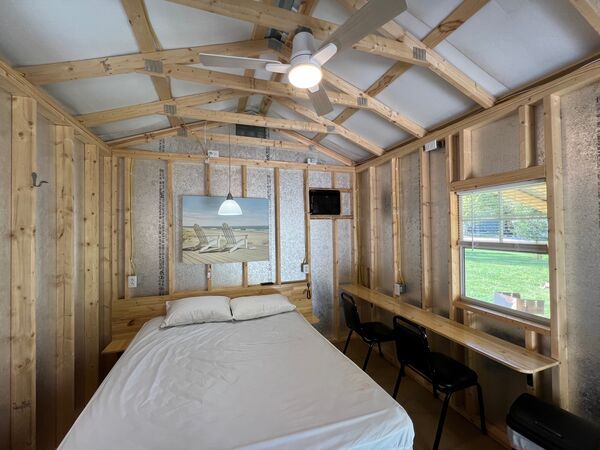 Queen bed with two pillows under a ceiling fan and a framed beach print, a long wooden counter with two chairs beneath the window