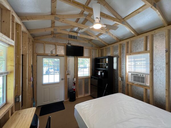 From the bed toward the front door: a mounted television, a black shelf unit with a mini fridge and microwave, and a window air conditioner