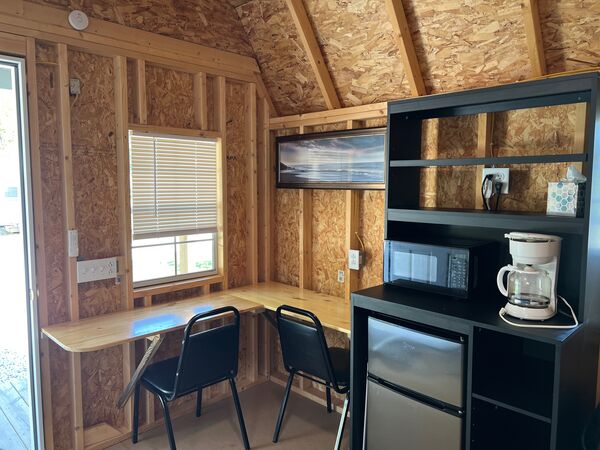 Kitchenette with a black shelf unit holding a microwave, coffee maker and mini fridge, beside a wooden counter with two chairs under a window