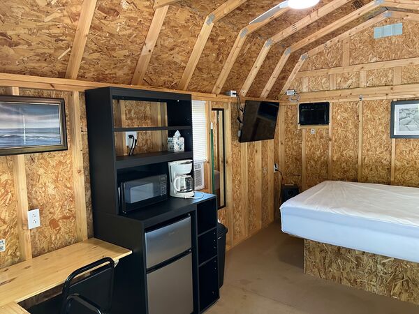 Interior with the kitchenette shelf unit and wall-mounted television, and a queen bed on a raised platform under the curved barn-style ceiling