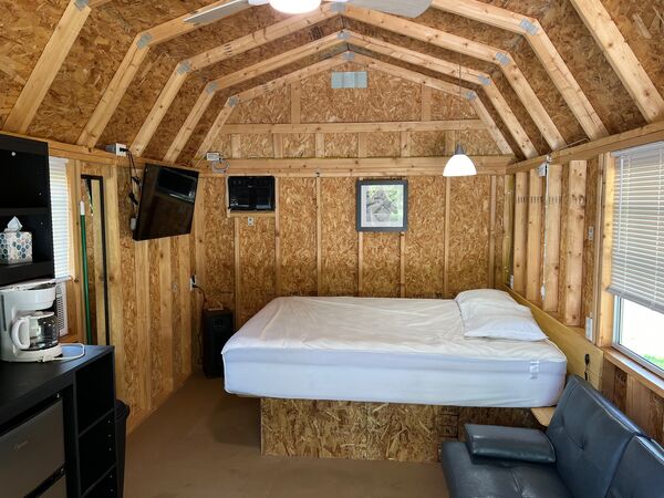 Queen bed on a platform at the end of the cabin under a pendant light, with a mounted television, a black fold-down couch and a coffee maker in view