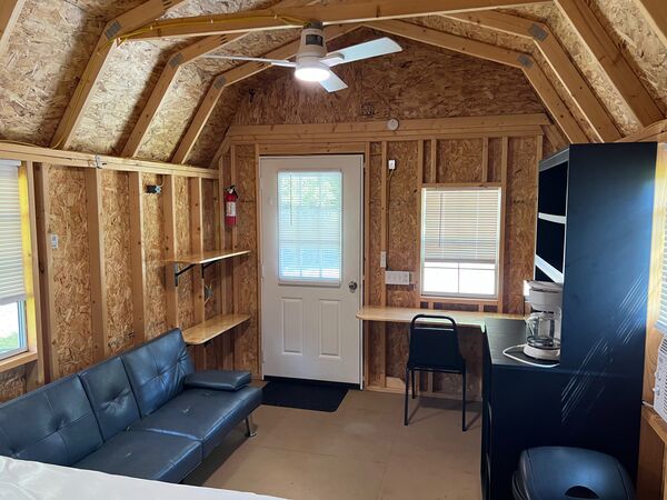 Sitting area with a black fold-down couch, wall shelves, the front door and a small desk with a chair, under a ceiling fan