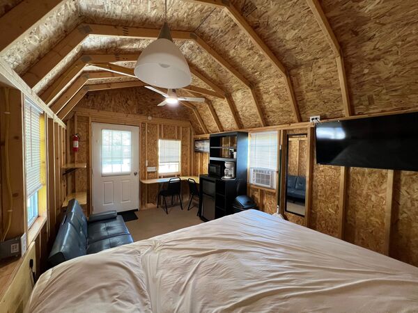 From the bed across the cabin: the fold-down couch, front door, a desk with chairs, the kitchenette shelf unit, a window air conditioner and a mounted television