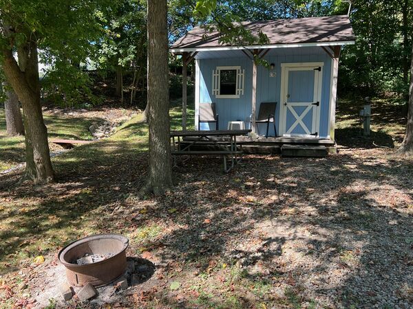 Light blue cabin in a wooded, shaded site with a picnic table in front of the porch and a fire ring in the foreground