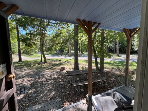 View from the porch under the roof to a picnic table, fire ring and grill on a shaded wooded site, with a gravel road beyond