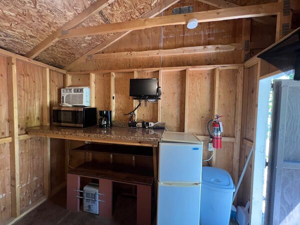 Kitchenette counter with a microwave, coffee maker and small television, a window air conditioner above and a white refrigerator and trash can beside it