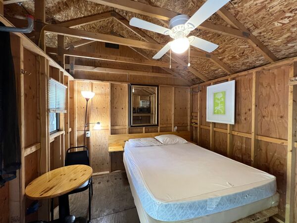 Queen bed under a ceiling fan with a mirror over the headboard, a green botanical print, a floor lamp and a small round table with a chair