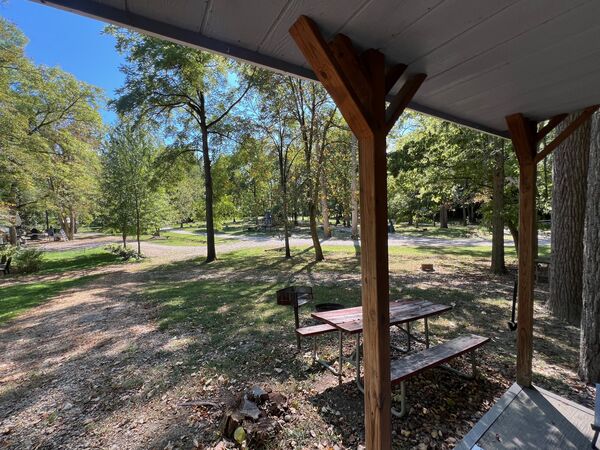 View from the porch to a picnic table and charcoal grill on a shaded site, with tall trees and a gravel road beyond