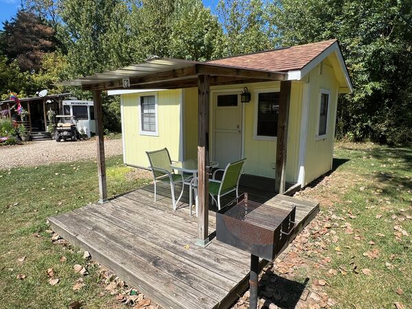 Covered front deck with two green chairs, a glass bistro table and a charcoal grill on a post