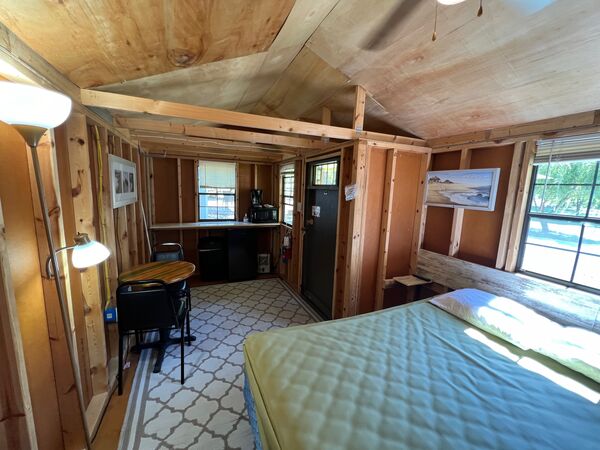 Inside the cabin: a queen bed by the window, a small round table and chair, a floor lamp, and a counter with a coffee maker and microwave at the far end