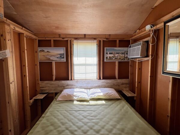 Queen bed beneath a window, framed beach prints on the wood walls on either side and a wall air conditioner