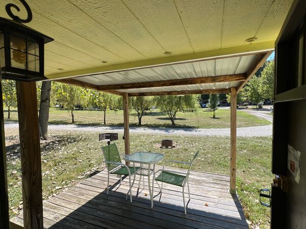 View from the doorway across the covered deck and its table and chairs to the lawn, fire ring, grill and a tree-lined gravel road