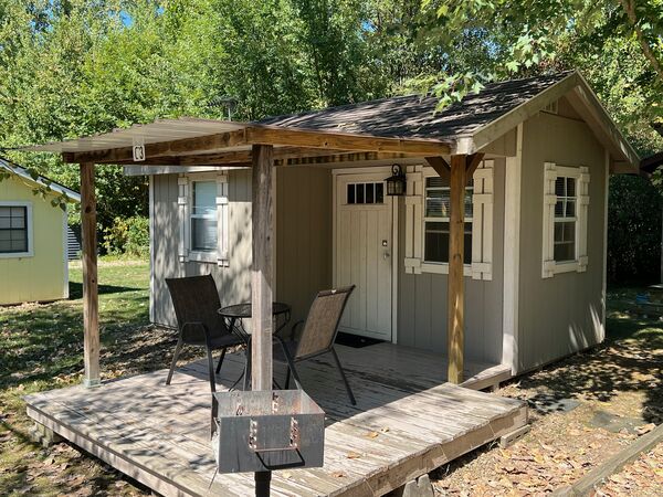 Covered front deck with two black mesh chairs, a small round table and a charcoal grill on a post beside the door