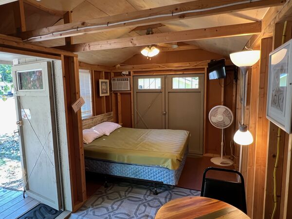 Interior from the front door: a queen bed with two pillows, a wall air conditioner, mounted television, ceiling fan, floor fan and floor lamp