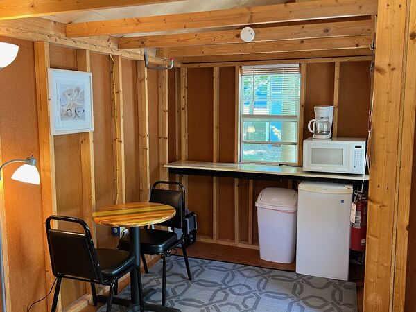 Kitchenette counter under a window with a coffee maker and microwave, a mini fridge and trash can below, beside a round table and two chairs