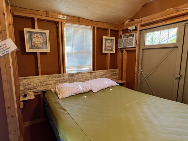 Queen bed against a rustic wood headboard with small shelves on each side, under a window, with a wall air conditioner by the double doors
