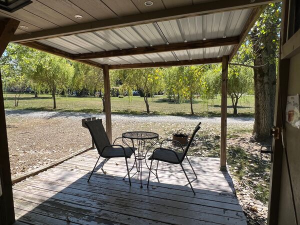 View from the doorway across the covered deck and its two chairs and table to a gravel road and a grassy field lined with willow trees