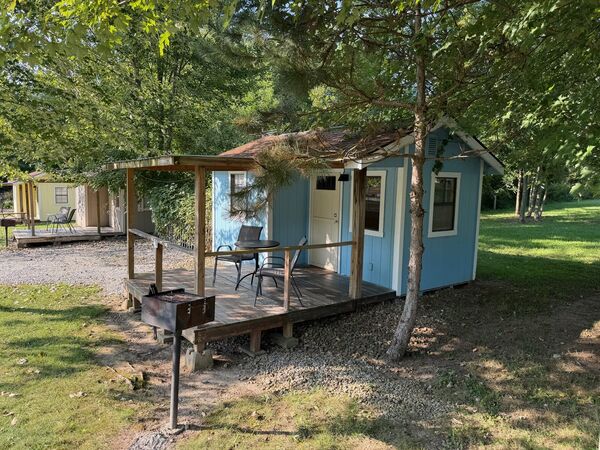 Light blue cabin seen from the side, its railed deck holding a table and chairs, with a charcoal grill in front and neighboring cabins beyond