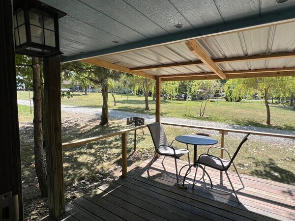 View from the doorway across the covered, railed deck with two chairs and a small table, to a gravel road and a field of willow trees