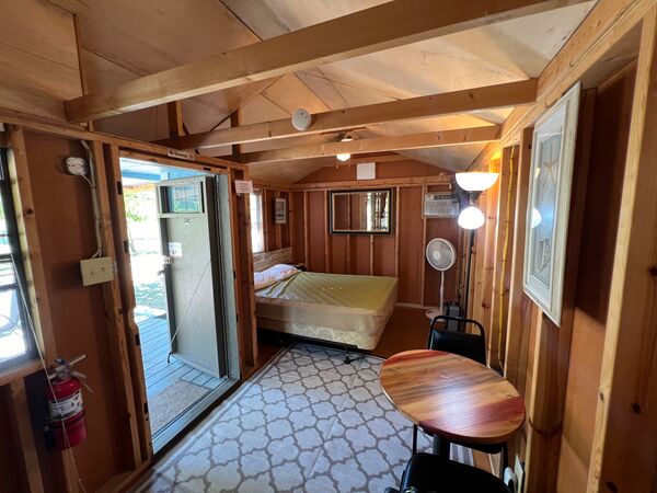 Interior from beside the door: a queen bed at the far end under a mirror and wall air conditioner, with a floor fan, floor lamp and a round table with chairs