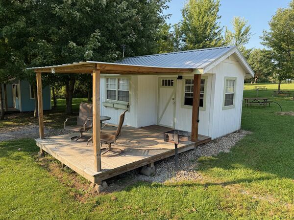 White cabin from the side, its covered deck with two chairs and a table and a charcoal grill beside it, picnic tables on the grass beyond
