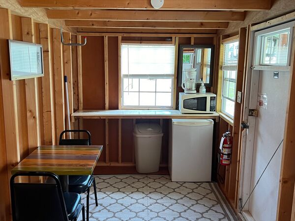 Kitchenette counter under a window with a coffee maker, microwave and mirror, a mini fridge and trash can below, beside a table with two chairs
