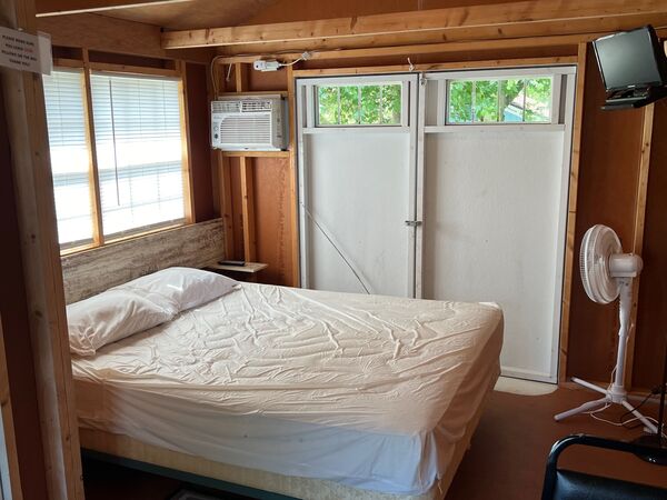 Queen bed against a rustic wood headboard under a window, with double doors, a wall air conditioner, a television on a wall arm and a floor fan