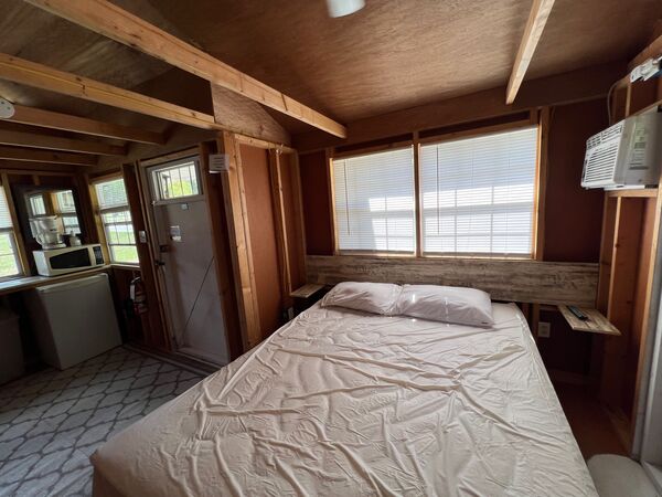 Queen bed beneath a pair of windows with a wall air conditioner, and the kitchenette, mini fridge and front door across the room