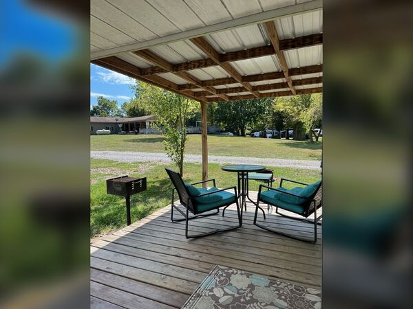 Covered deck with two teal-cushioned rocking chairs, a small table, an outdoor rug and a charcoal grill, overlooking a grassy field