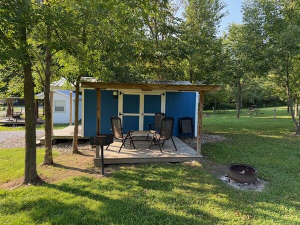 Covered porch outside blue double doors with folding chairs and a small table, a charcoal grill at the edge and a fire ring on the lawn