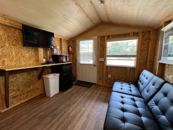 Sitting area with a black fold-down couch, a counter with a mounted television, microwave and coffee maker, and a mini fridge beside the front door