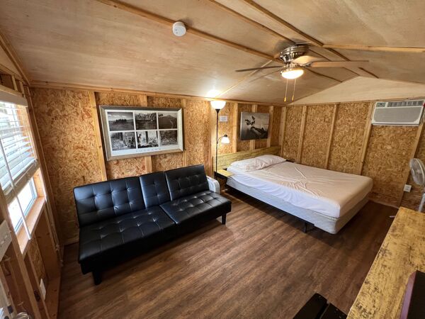 Interior with a queen bed against the far wall, a black fold-down couch, a framed black-and-white photo, a ceiling fan and a wall air conditioner