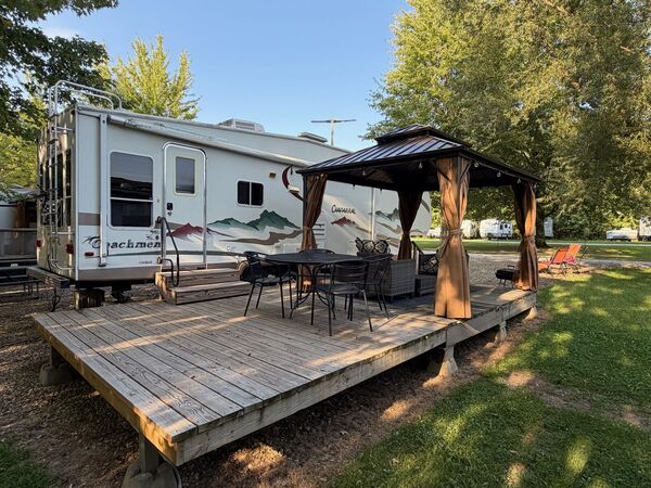 Large wooden deck along the trailer with a curtained gazebo over a dining table and chairs, and lounge chairs on the lawn beyond
