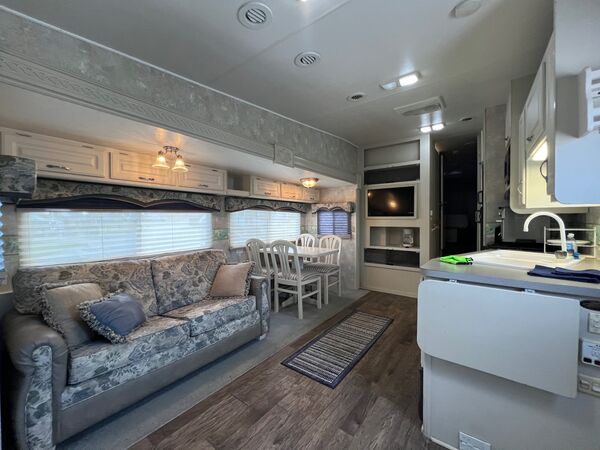 Living area with a floral sofa in the slide-out, a dining table with four chairs, a television in the cabinetry and the kitchen sink