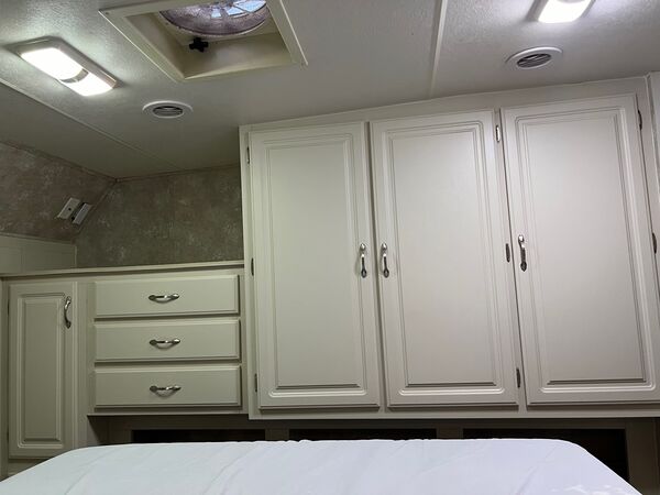 Bedroom wall of white built-in closets and drawers under a roof vent, seen over the foot of the bed