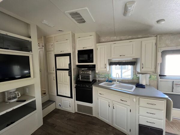 Kitchen with white cabinets, a sink under a window, a propane stove with a microwave above, an air fryer oven and a refrigerator, beside the television nook
