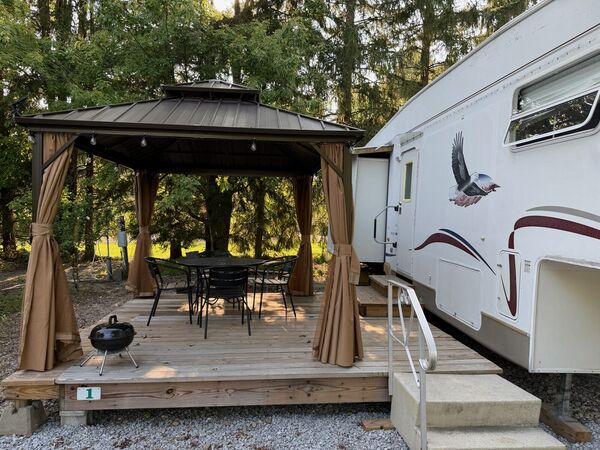 Deck with a curtained gazebo sheltering a dining table and chairs, a small kettle grill, and steps with a handrail up to the trailer door
