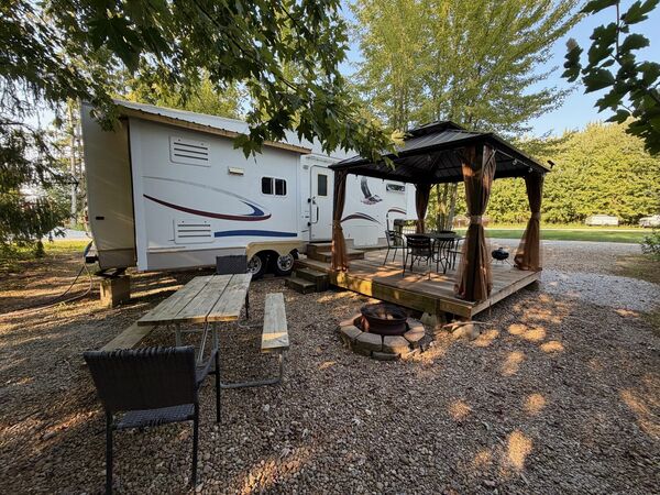 Trailer side with the gazebo deck, a picnic table, a wicker chair and a stone-ringed fire pit on the gravel site